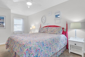 Quarto com cama, mesa de cabeceira, abajur, janela com persianas e relógio de parede.