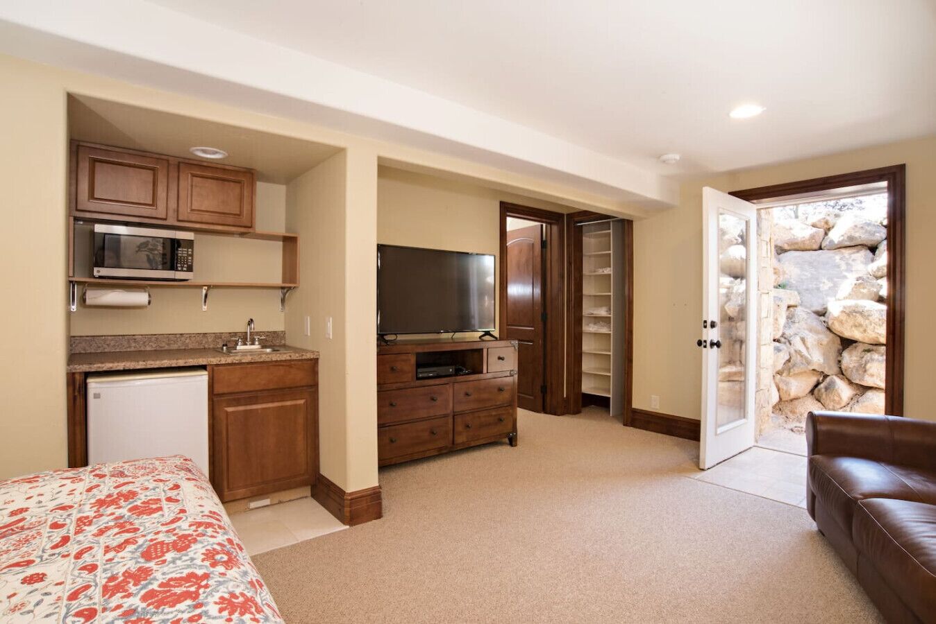 A compact room with a kitchenette, a bed with floral bedding, a television, and a stone wall.