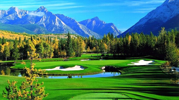 A golf course with a pond, surrounded by autumn-colored trees and mountains.