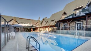 A swimming pool with clear water, surrounded by stone buildings with large windows and a covered area with chairs.