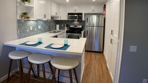 Una cocina moderna con gabinetes blancos, un refrigerador de acero inoxidable, un microondas y un lavamanos. Hay tres taburetes en la isla y una planta en maceta sobre la encimera.