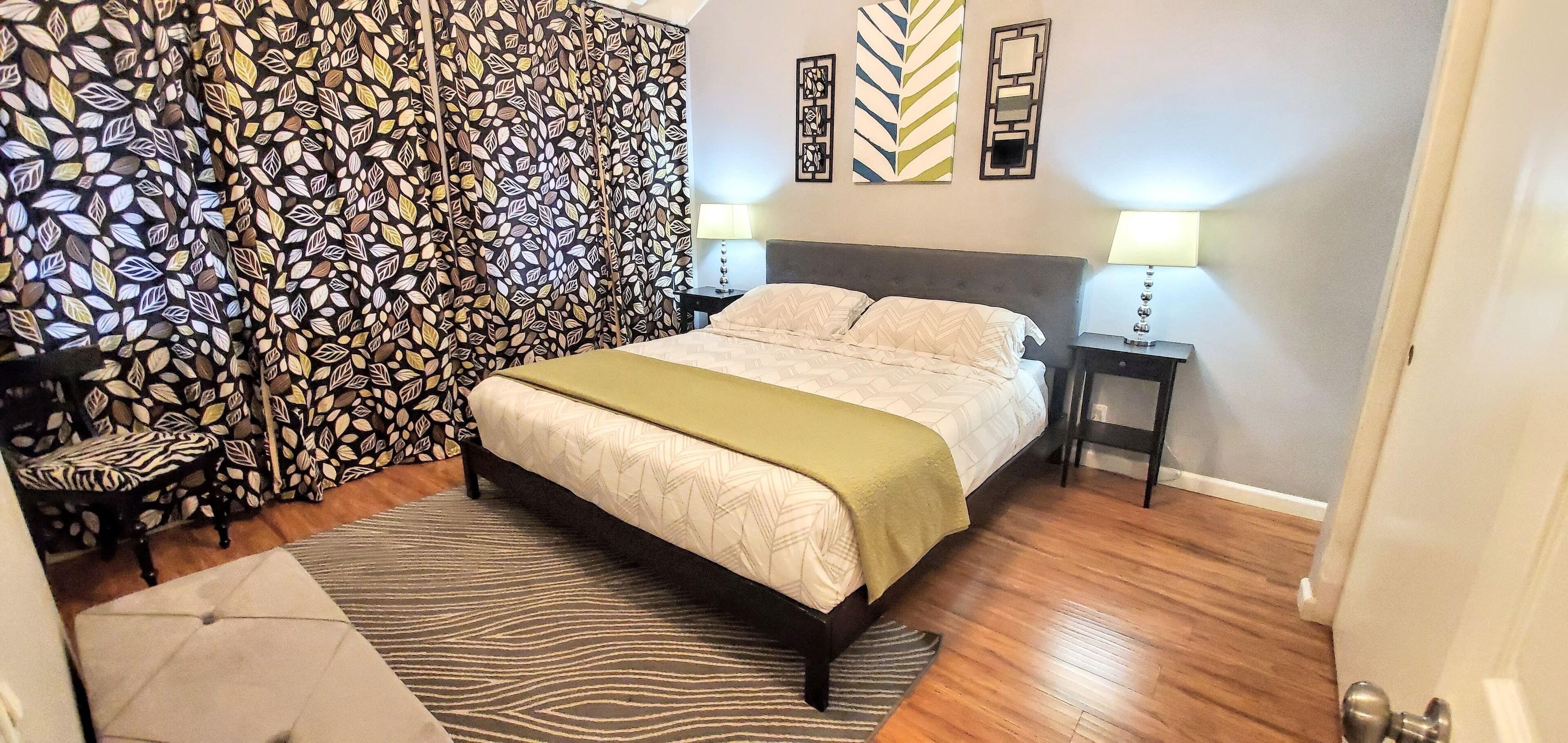 Un dormitorio con cama, cortina estampada, mesita de noche con lámpara y pared decorada con paneles.