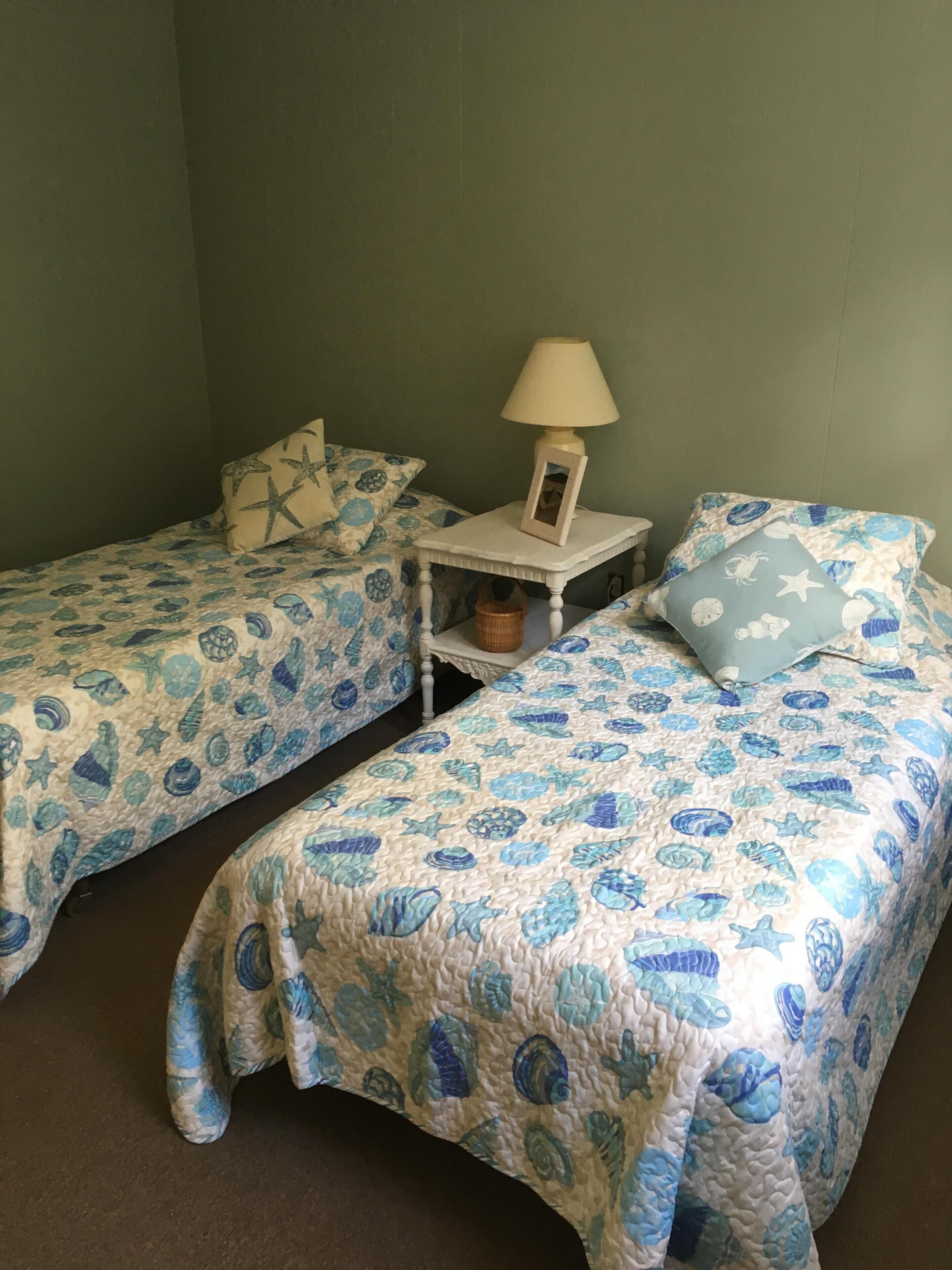 Een kamer met twee bedden, elk met een blauw-wit gebed, een klein nachtkastje met een lamp, en een wandplank met een mand.