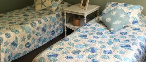 Een kamer met twee bedden, elk met een blauw-wit gebed, een klein nachtkastje met een lamp, en een wandplank met een mand.
