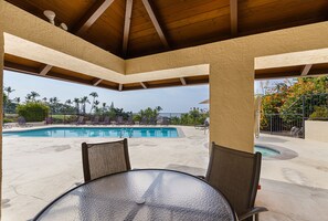 A covered outdoor pool area with lounge chairs and a table, overlooking a swimming pool and palm trees.