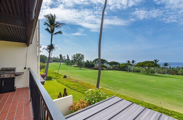 A balcony with a grill, overlooking a well-manicured lawn and palm trees, with a clear view of the ocean.