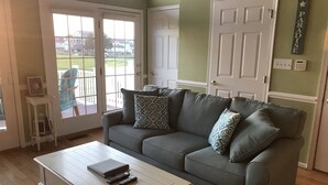 Ein Wohnzimmer mit grauem Sofa, gemusterten Kissen, einem weißen Couchtisch mit Fernbedienungen und einer Tür mit dem Schild 'PARADISE'.