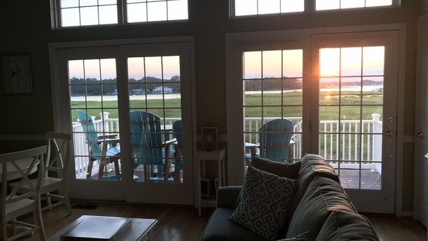 Ein Wohnzimmer mit einer Couch, Sesseln und einem Tisch. Zwei große Fenster bieten Blick auf eine Rasenfläche und ein Gebäude.