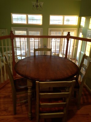 Un comedor con una mesa redonda de madera rodeada de sillas, ventanas amplias con persianas y una escalera con barandilla.