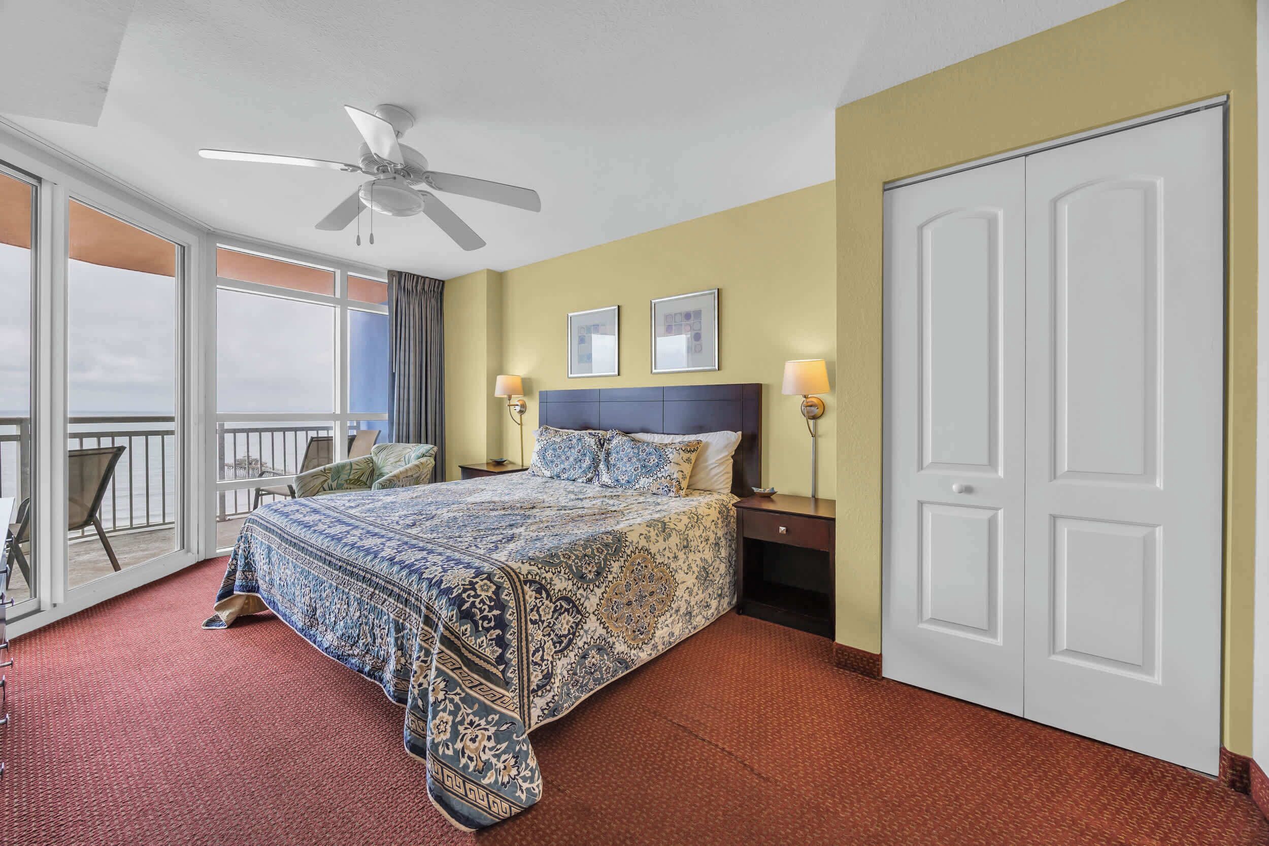 A hotel room with a large bed, a ceiling fan, a balcony with a chair, and a white door.