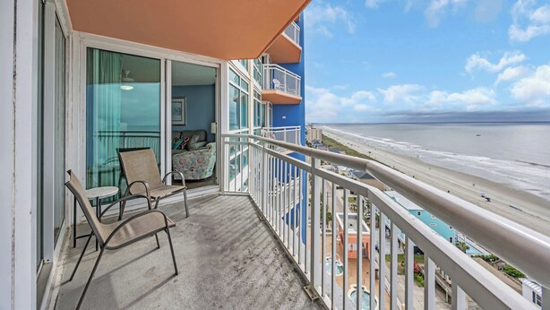 A balcony with a view of the beach, a pool, and a building with multiple balconies. - Prince Luxury Ocean View w Sky Pool Lazy River (North Myrtle Beach)