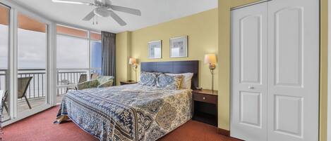 A hotel room with a large bed, a ceiling fan, a balcony with a chair, and a white door.