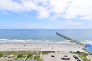 A beach with a pier extending into the water, a parking area, and a clear sky.