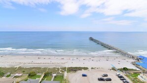 A beach with a pier extending into the ocean, a parking lot, and a few buildings in the foreground. - Prince Luxury Ocean View w Sky Pool Lazy River (North Myrtle Beach)