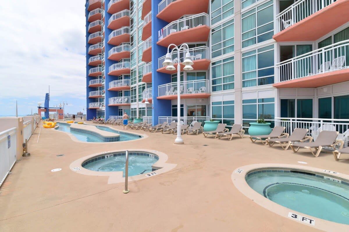 A cruise ship deck with multiple swimming pools, lounge chairs, and a large building with balconies.