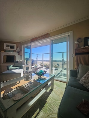 A living room with a glass sliding door leading to a deck, a fireplace, and a view of the beach.
