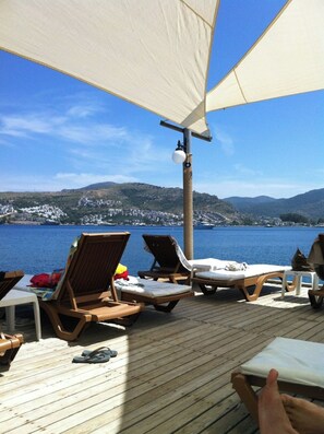 Een houten terras met lounge stoelen en een tafel, met uitzicht op een waterpartij en heuvels op de achtergrond.