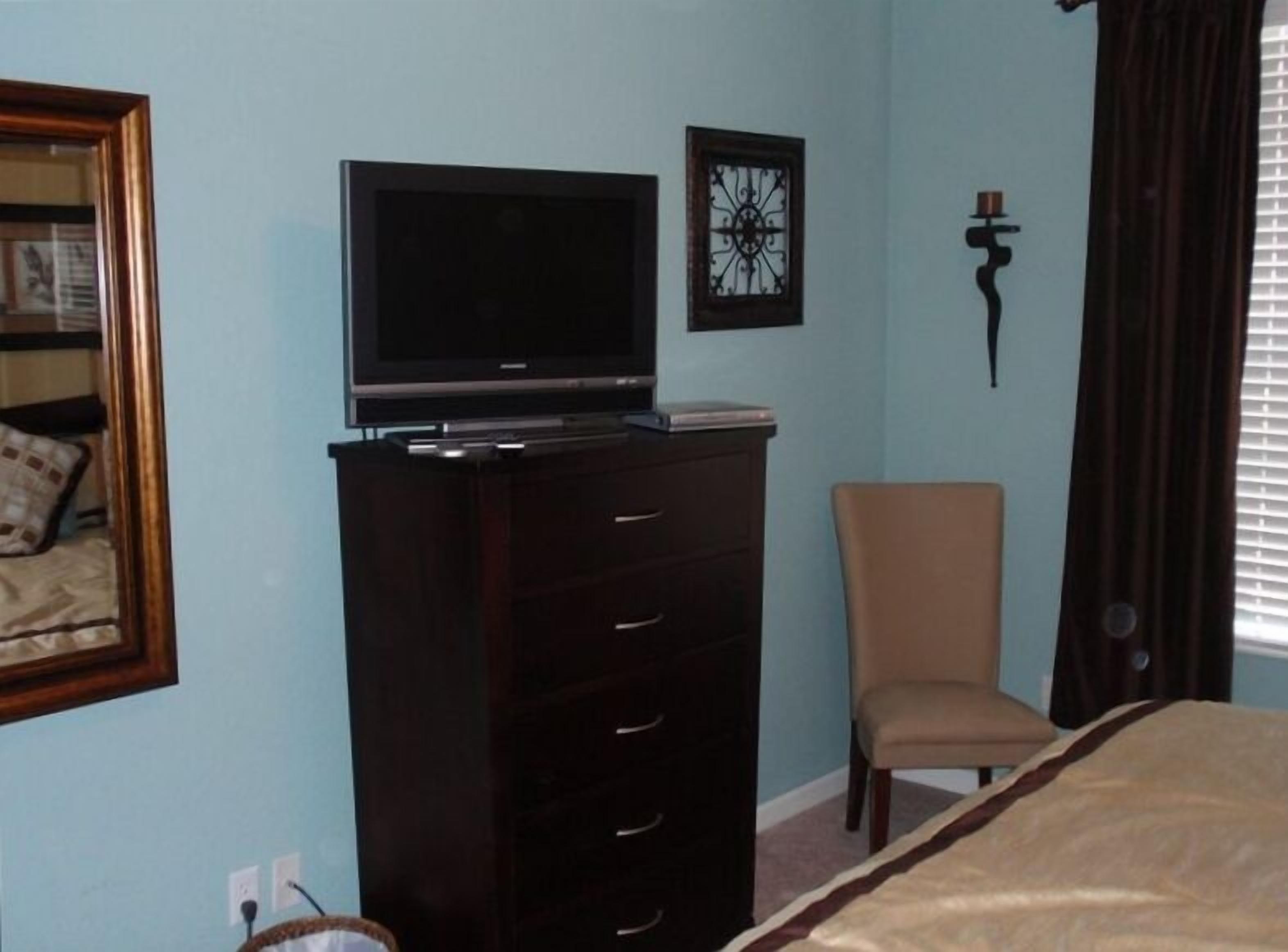 A bedroom with a bed, a dresser, a chair, a TV on the dresser, and a framed picture on the wall.