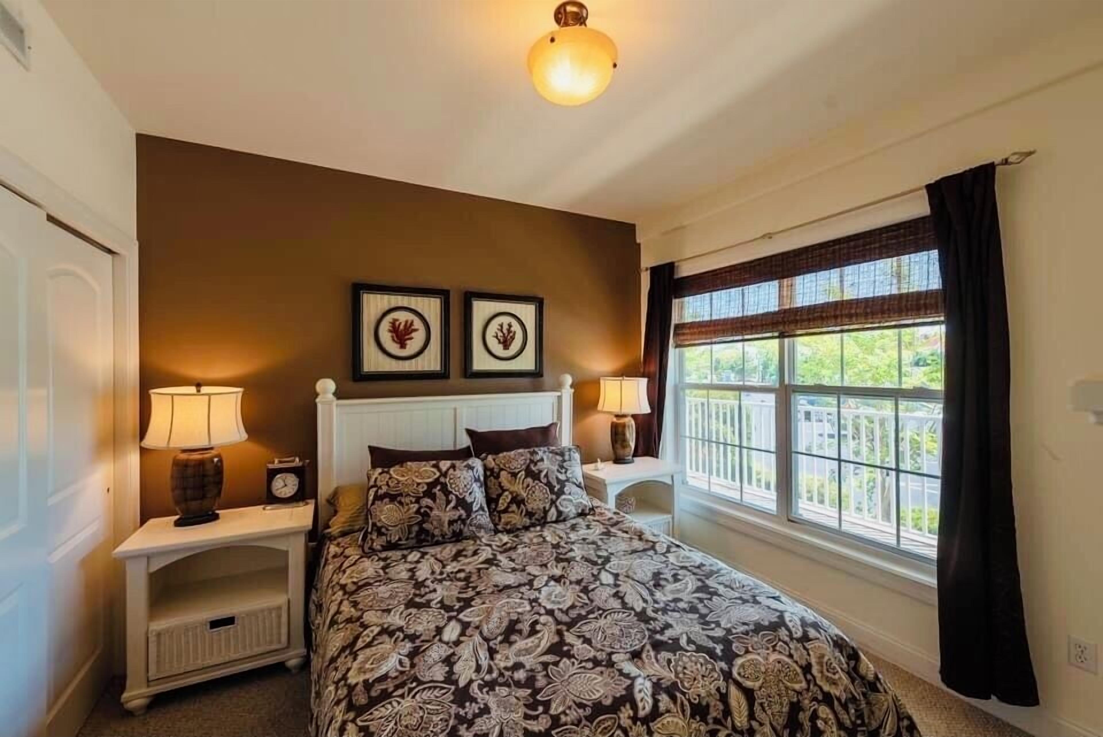 Un dormitorio con cama, dos mesitas de noche con lámparas, una ventana con cortinas y dos cuadros colgados en la pared.