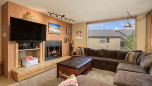 A living room with a fireplace, a large flat-screen TV, a brown leather couch, and a window with a view of a house.