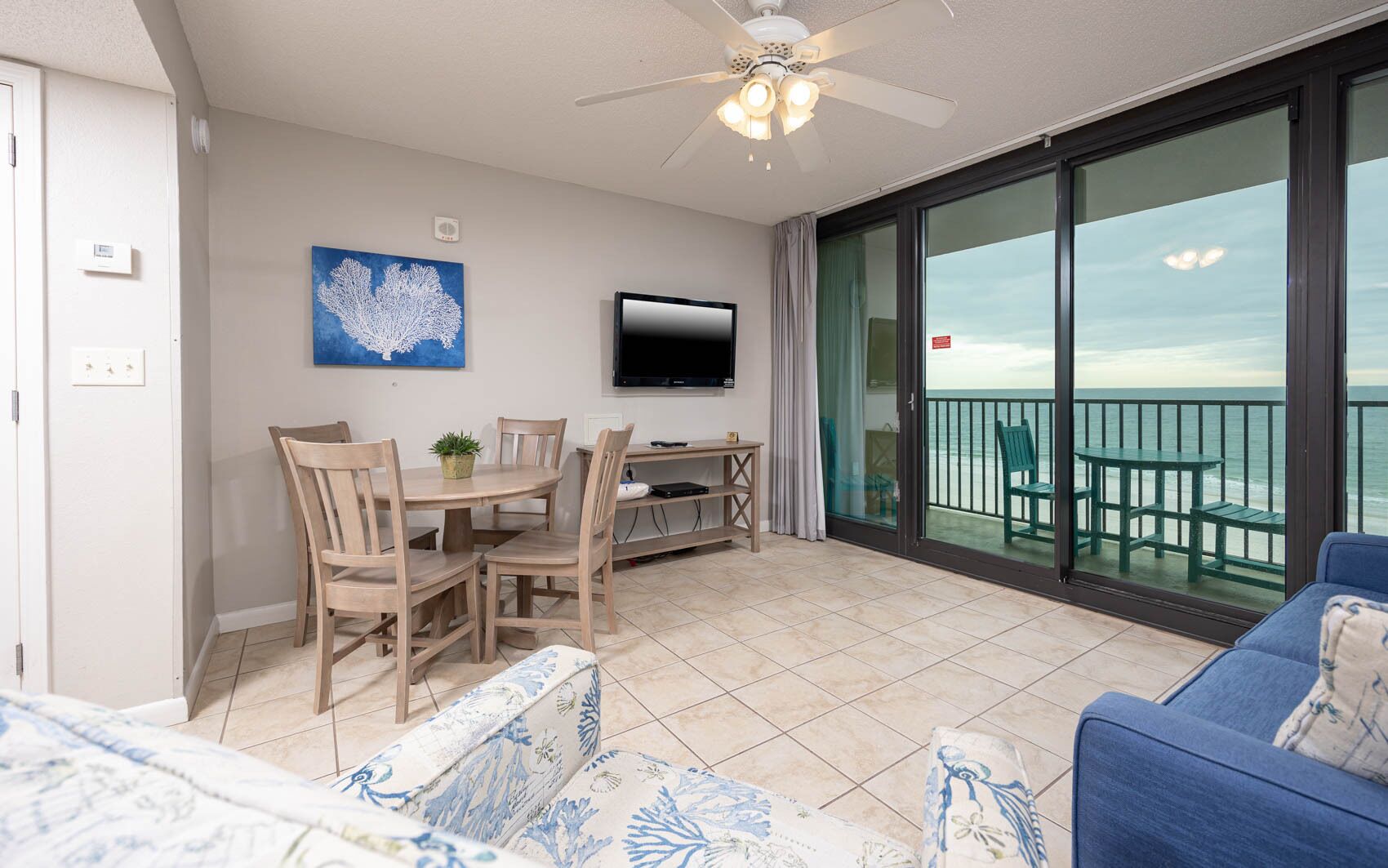 Un balcon avec vue sur la mer, un coin repas avec des chaises et une table, un téléviseur fixé au mur et un ventilateur de plafond.
