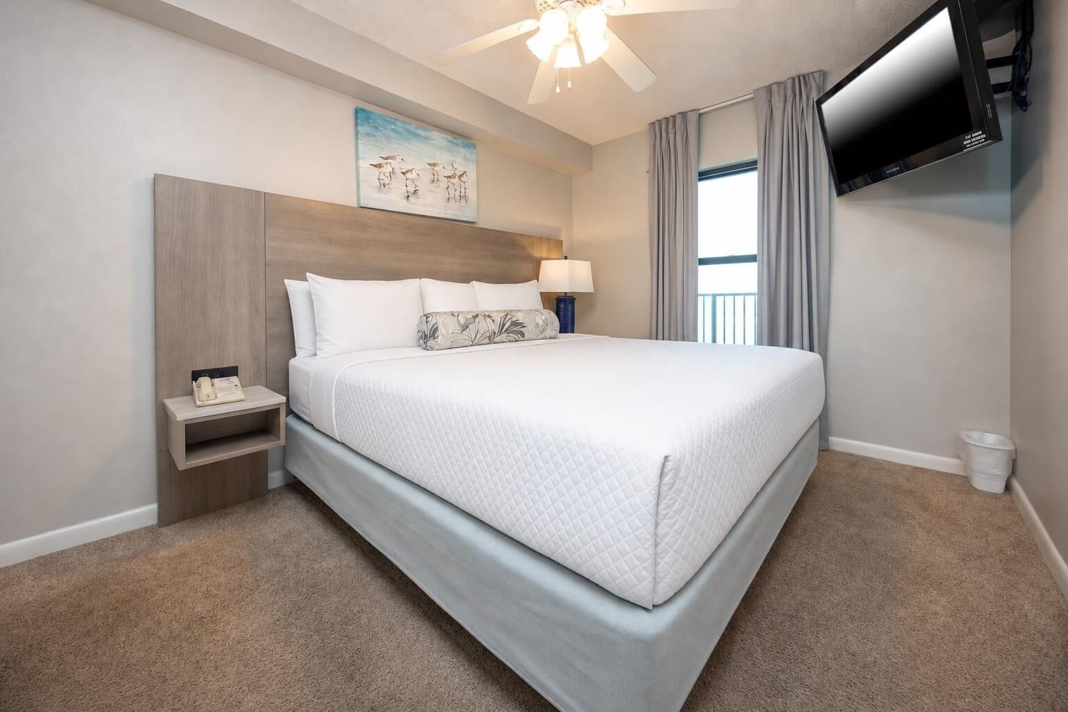 Une chambre d’hôtel avec un lit, une table de chevet, une lampe, un ventilateur de plafond, une télévision et un tableau au mur.