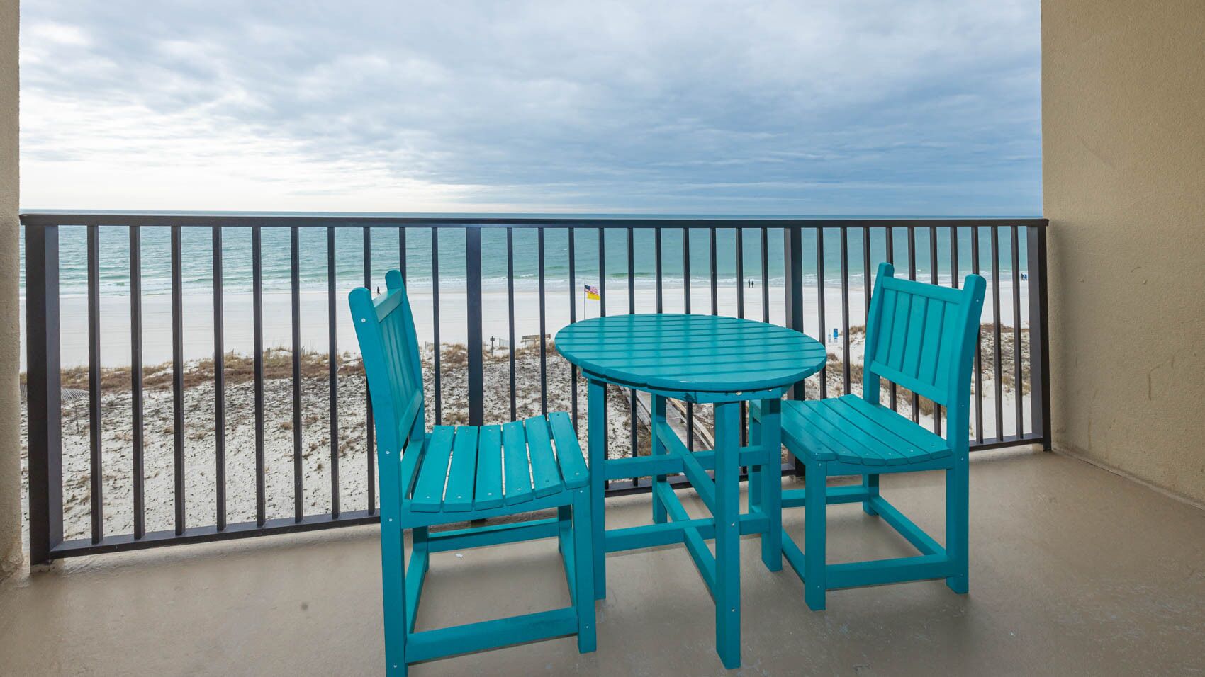 A balcony with a view of the beach, featuring two turquoise chairs and a small round table.