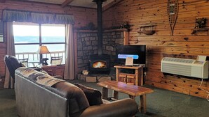 Ein gemütliches Blockhaus-Interieur mit Ledersofa, Kamin aus Stein, Fernseher und Blick ins Freie.