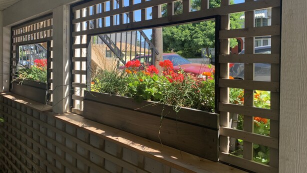 Une fenêtre ornée d’un treillis en bois, à travers laquelle on aperçoit une voiture rouge et de la verdure.