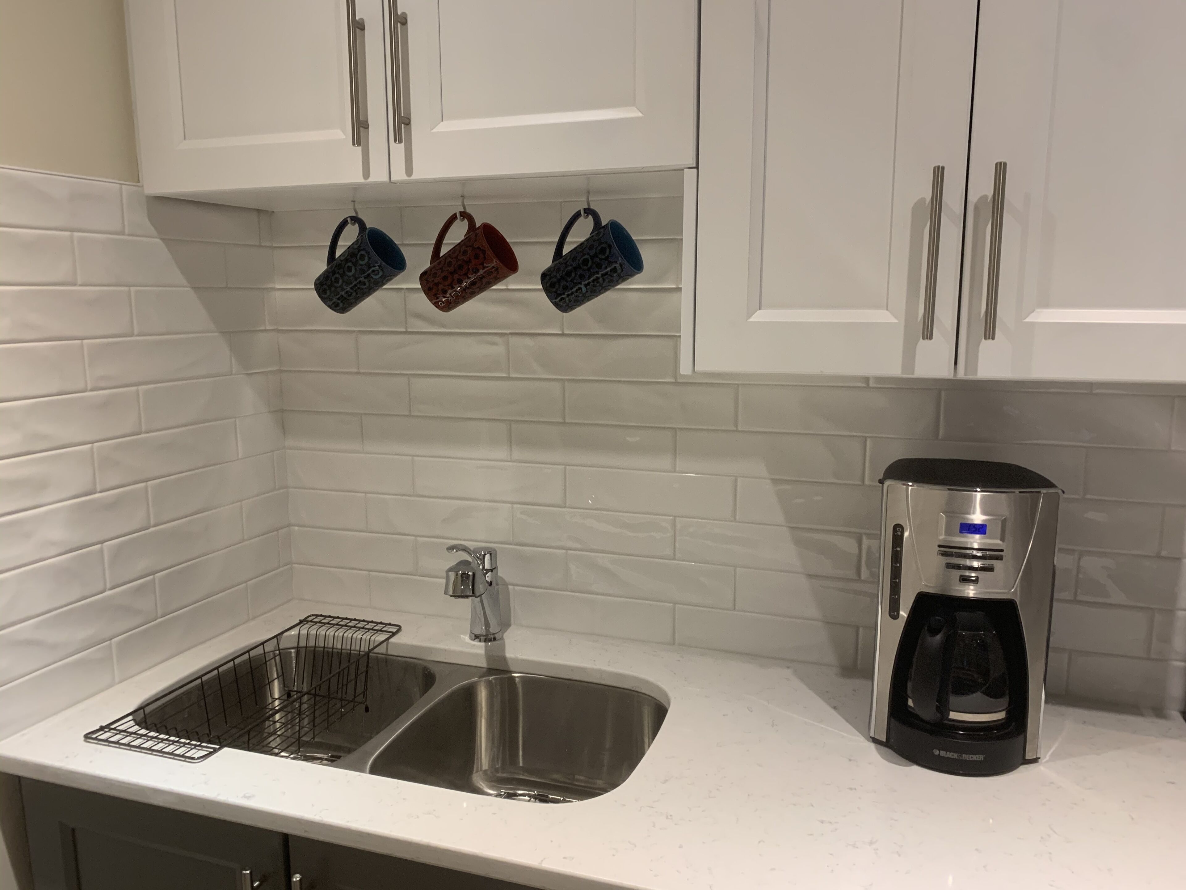 Een keuken met witte kastjes, een roestvrijstalen spoelbak, een koffiezetapparaat en drie hangende mokken.