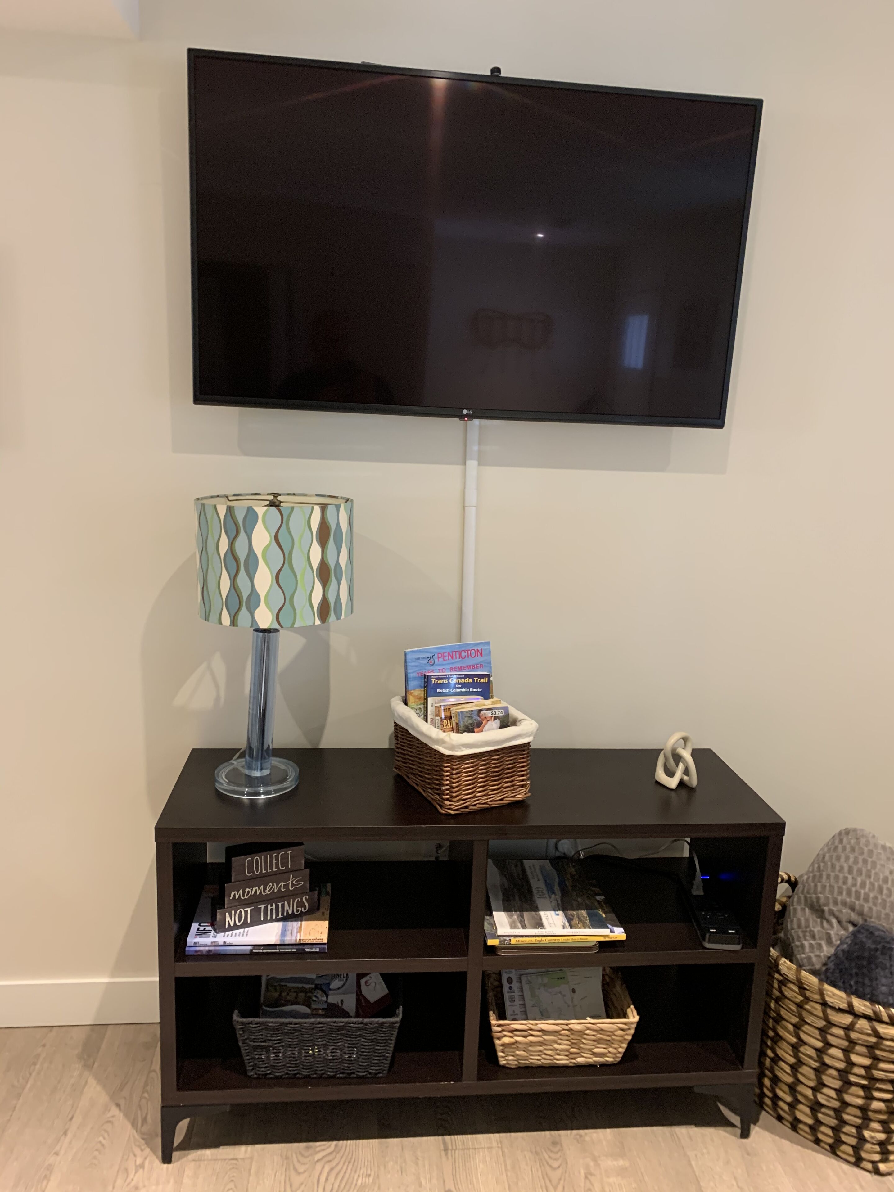 A dark wooden TV stand with a flat-screen TV mounted above it. On the stand, there is a lamp with a patterned lampshade, a basket containing a book titled "PENTICOTON" and a map, and a small decorative item.