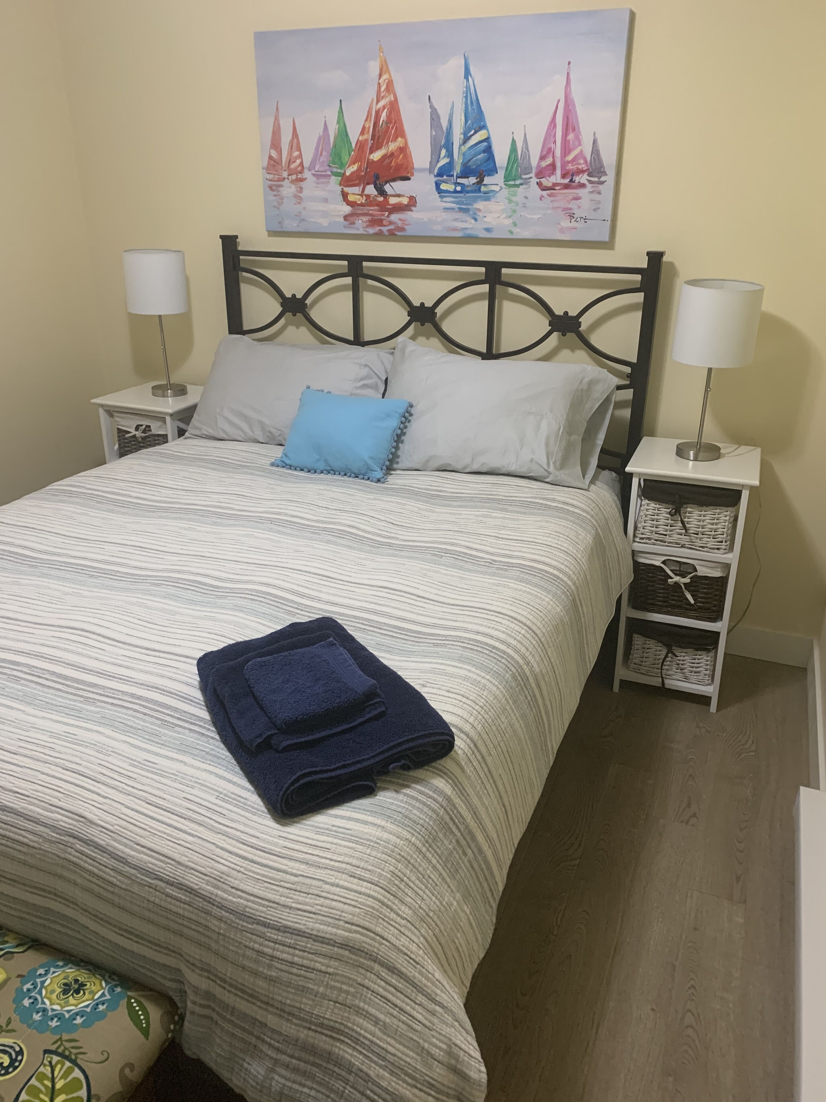 A neatly made bed with a striped bedspread, two pillows, and a folded blue towel. There are two bedside tables with lamps and a wall-mounted painting of sailboats.