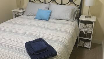 Un lit bien fait, recouvert d’une courtepointe à rayures, avec deux oreillers et une serviette bleue pliée. On y trouve deux tables de chevet avec des lampes et, au mur, un tableau représentant des voiliers.