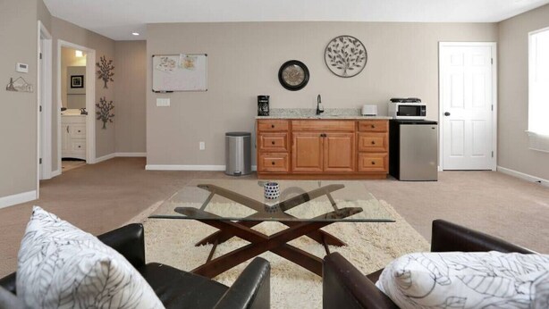 A modern living room with a glass coffee table, two leather armchairs, a kitchen with wooden cabinets, a refrigerator, and a white door.