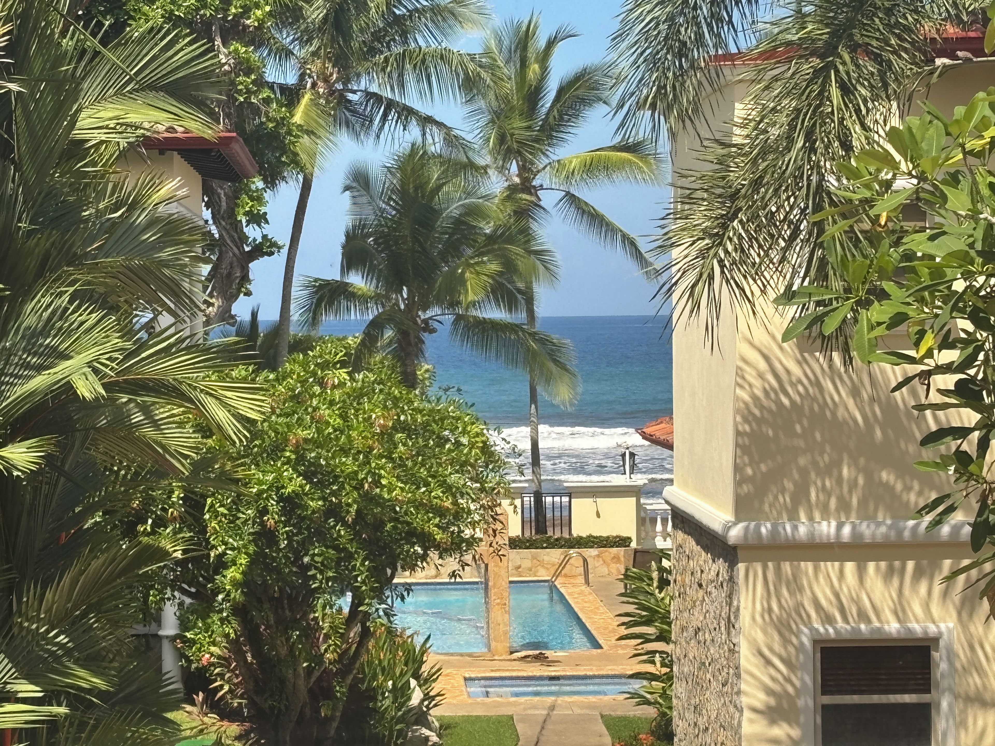 A pool area with a view of the ocean, surrounded by palm trees and a building with a stone facade.