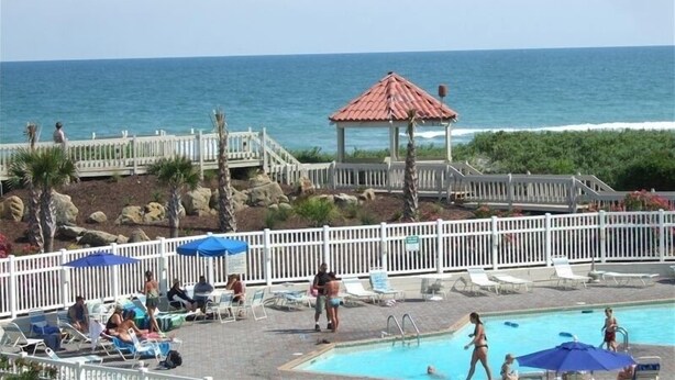 A pool area with lounge chairs and umbrellas, a gazebo, and a view of the ocean. - Perfect condo for your getaway.  (N Topsail Beach)