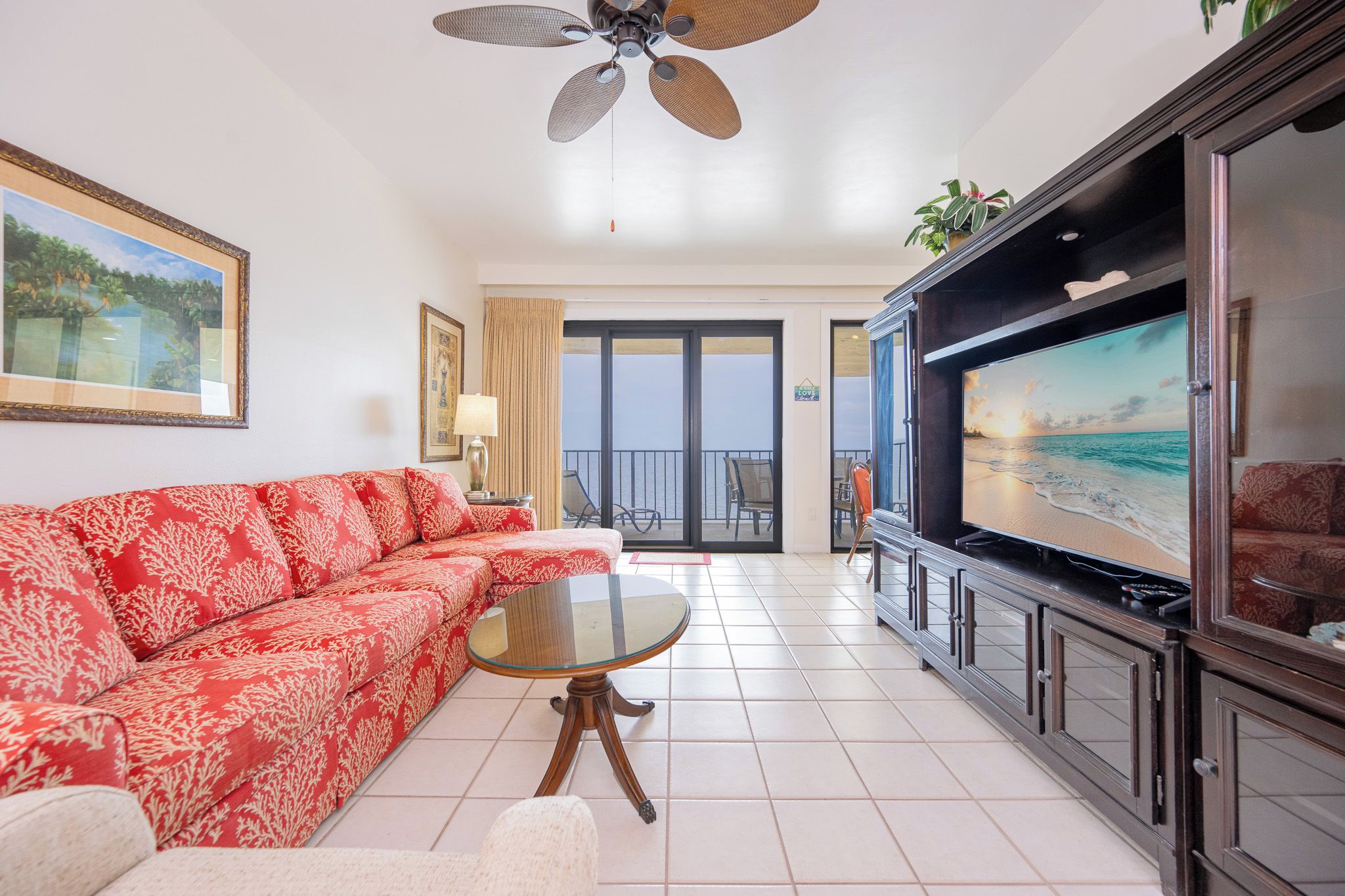 Ein Wohnzimmer mit einem roten Sofa, einem Glastisch, einem Deckenventilator, einem Fernseher und einem Balkon mit Meerblick.