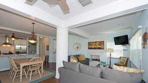 Una sala de estar con un sofá gris, una mesa de comedor con sillas, una zona de cocina con refrigerador y un televisor montado en la pared.