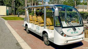 A beach trolley with a covered seating area, parked on a paved path with palm trees and buildings in the background.