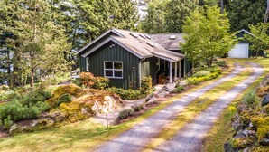 A green wooden cabin with a porch, surrounded by lush greenery and a gravel path. - Orcas Island, Casa Verde #269 (Eastsound)