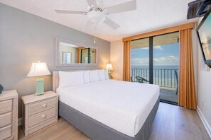Een hotelkamer met een bed, nachtkastjes, een plafondventilator, een groot raam met uitzicht op zee en een balkon met een stoel.