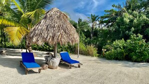 Una playa con dos sillas azules de descanso bajo un toldo de paja, rodeada de palmeras y follaje.