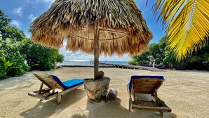 Una playa con dos sillones de madera bajo un paraguas de paja, rodeada de palmeras y una vista despejada del océano.