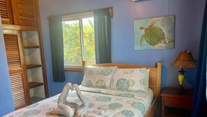 Una cama bien hecha con un arreglo de almohadas con forma de cisne, una ventana con cortinas, un cuadro de una tortuga marina y una lámpara de noche.