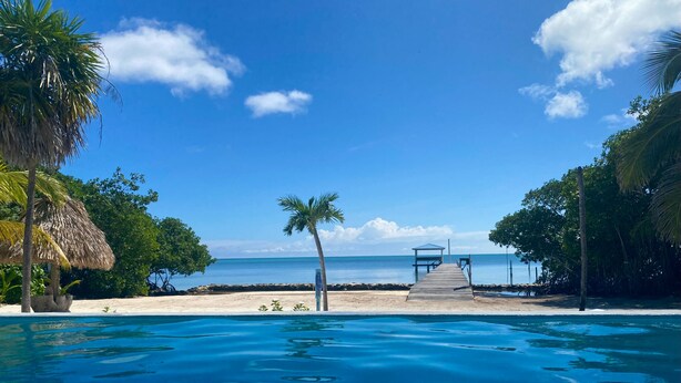 Una piscina azul cristalina con una terraza de madera que da a la playa.