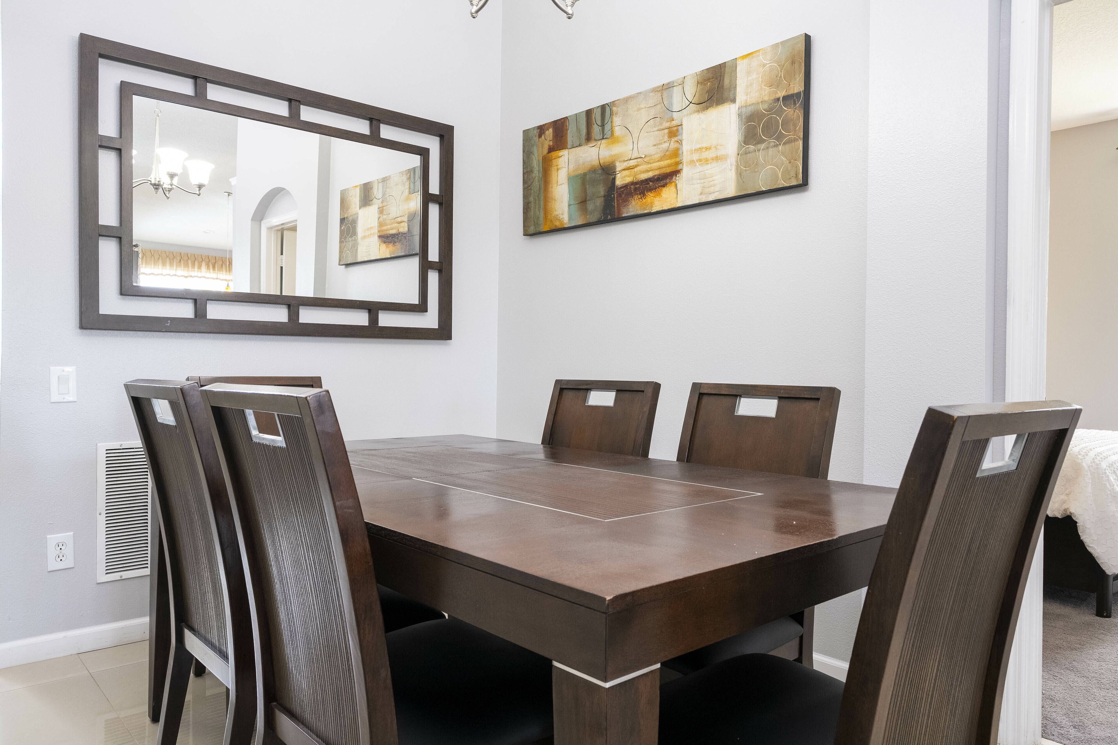Een eethoek met een rechthoekige tafel en zes stoelen, een grote spiegel en een modern abstract schilderij aan de muur.