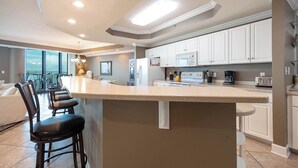 A modern kitchen with white cabinets, a central island with bar stools, and a view to a balcony. - Bay Breezes, Perfect Ease Lighthouse on the Bay 2513 (Orange Beach)