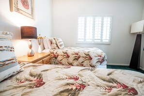 Une chambre à coucher avec un lit, des tables de chevet, une lampe, une fenêtre avec des stores et un tableau encadré au mur.