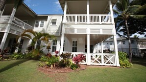A two-story white house with a balcony, surrounded by tropical plants and palm trees.