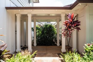 A covered porch with a concrete pathway leading to a door, surrounded by various plants and a large flowering plant on the right.
